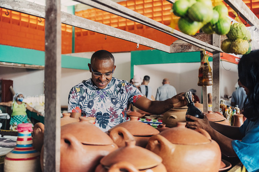 smiling young market vendor behind his pottery stall showing his products to female client at public market in Bahia, Brazil