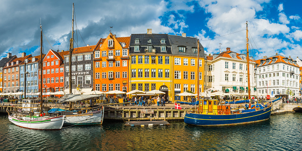 Crowds of people sitting, walking, chatting and relaxing on the picturesque waterfront of Nyhavn, its brightly coloured historic houses, cafes, bars, restaurants and nightclubs reflecting in the tranquil blue waters of the harbour canal leading to Kongens Nytor.