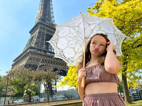 beautiful young teenager girl in paris on the background of the eiffel tower in a long elegant dress in the style of romanticism walks with an umbrella from the sun and smiles at her long blonde hair and around good weather and in the background the eiffe.