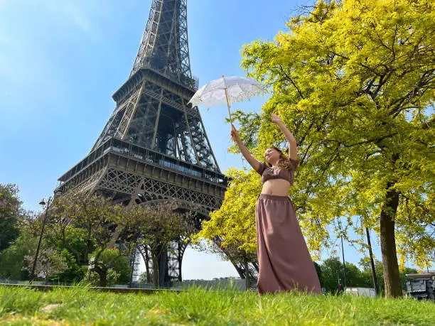 Photo of beautiful young teenager girl in paris on the background of the eiffel tower in a long elegant dress in the style of romanticism walks with an umbrella from the sun and smiles at her long blonde hair and around good weather and in the background the eiffe
