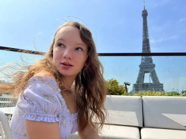 Photo of beautiful young teenager girl in paris on the background of the eiffel tower in a long elegant dress in the style of romanticism walks with an umbrella from the sun and smiles at her long blonde hair and around good weather and in the background the eiffe