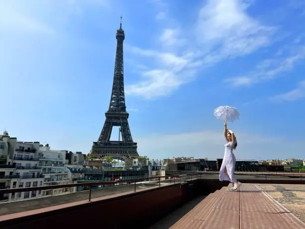 Photo of beautiful young teenager girl in paris on the background of the eiffel tower in a long elegant dress in the style of romanticism walks with an umbrella from the sun and smiles at her long blonde hair and around good weather and in the background the eiffe