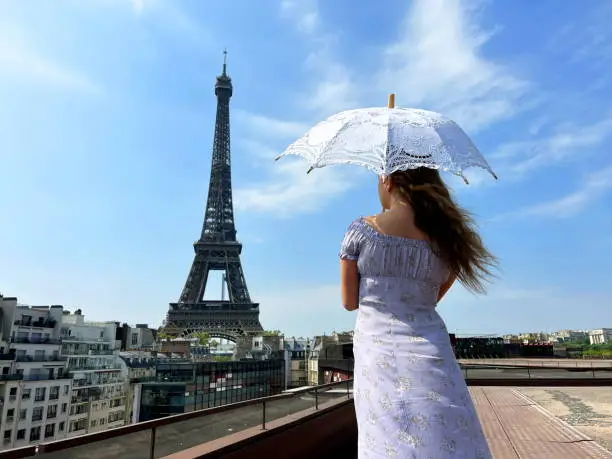 Photo of beautiful young teenager girl in paris on the background of the eiffel tower in a long elegant dress in the style of romanticism walks with an umbrella from the sun and smiles at her long blonde hair and around good weather and in the background the eiffe