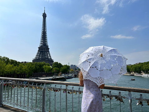 beautiful young teenager girl in paris on the background of the eiffel tower in a long elegant dress in the style of romanticism walks with an umbrella from the sun and smiles at her long blonde hair and around good weather and in the background the eiffe.