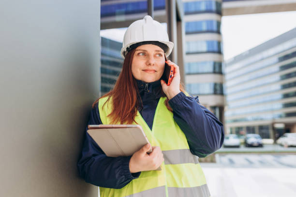 istock Happy Architect with a blueprints using smartphone at a construction site. Portrait of redhead woman constructor wearing white helmet and safety vest outdoors, technology or building concept 1402729955