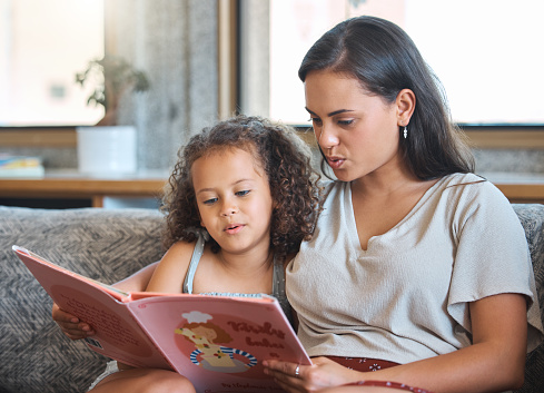 Loving hispanic mother and her little daughter sitting at home and reading a storybook together. Mother teaching little girl to read while sitting on the couch at home