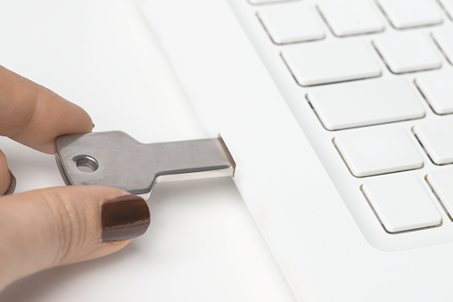 Female hand with red nail polish is holding silver colored USB stick attached to laptop on white background.