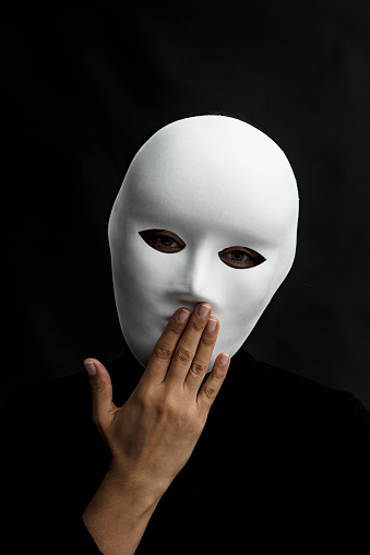 Unrecognizable female  is holding her hand in front her mouth and is wearing a white mask  in front of black background. She has to be quite and do not know what to say.