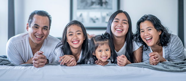 Three young Filipino sisters run into their parents bedroom and crash their parents bed, on a lazy Saturday morning. The mother and father are laying on their stomachs propped up on their elbows at the foot of the bed, with their hands folded out in front of them. They are laughing as their children stack in on top of them. They are all wearing their pajamas and laughing.