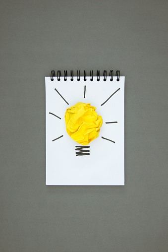 Light bulb made of yellow paper on white notepad on gray background.
