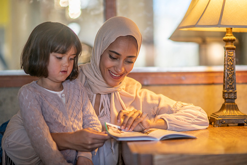 A young Muslim mother sits at a desk with her daughter as they read together under the lamp light. The little girl is seated o her mothers lap as she wraps her arms around her and hold the book out in front of them. They are both dressed casually and the mother is wearing a Hijab.