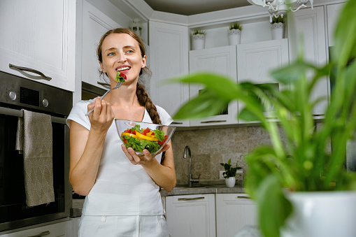 Young and happy woman eating healthy salad with green fresh ingredients from a bowl on the kitchen