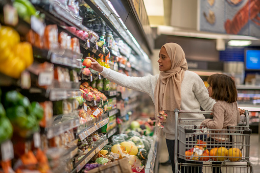 A young Muslim mother and daughter walk the produce aisles of the grocery store together shopping for groceries. They are both dressed casually and the mother is wearing a Hijab. The mother is pushing a shopping cart with her daughter in it and browsing the aisles.