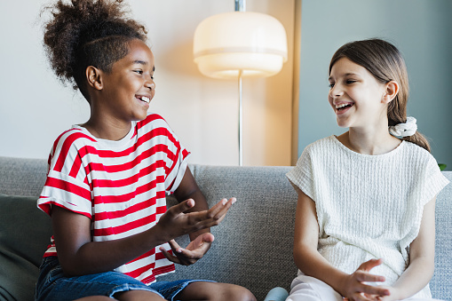 Two cute teenage girls are sitting on the couch at home. They are talking and laughing.