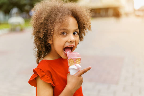 A black child in a red dress eats ice cream on a hot day. Ice cream in a waffle cone. A happy and contented child at summer.