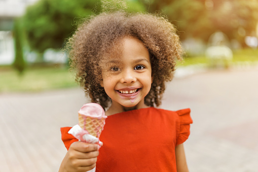 A black smileing child in a red dress eats ice cream on a hot summer day. Ice cream in a waffle cone. A happy and contented child at summer.