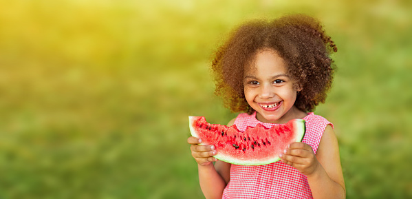 Funny Black kid eating watermelon outdoors in hot summer. Laughing baby looking at camera, healthy food