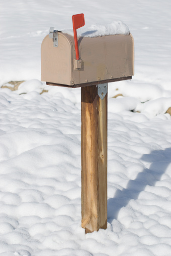 A beige rural mailbox with its red flag raised. It sits on a wooden post in fresh snow. There is snow on top of the box, too.