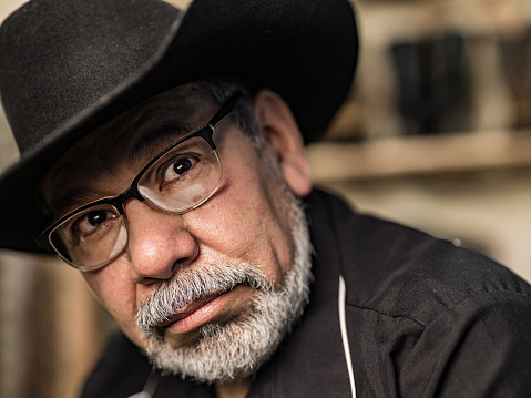 Portrait of mature Latin man dressed as Vaquero-Mexican Cowboy. He has grey hair beard, wearing eyeglasses and western themed clothes with cowboy hat, shirt and jeans. Interior of private home.