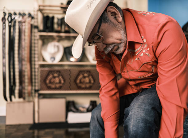 Mature Latin man getting dressed as Vaquero-Mexican Cowboy. He has grey hair beard, wearing eyeglasses and western themed clothes with cowboy hat, shirt and jeans. Interior of private home.