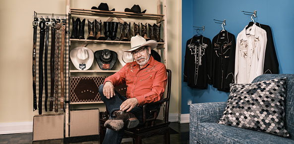 Portrait of mature Latin man dressed as Vaquero-Mexican Cowboy. He has grey hair beard, wearing eyeglasses and western themed clothes with cowboy hat, shirt and jeans. Interior of private home.
