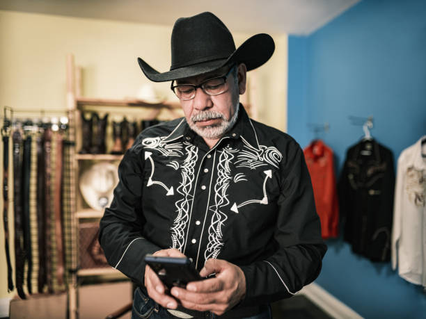 Mature Latin man dressed as Mexican Cowboy using mobile phone. He has grey hair beard, wearing eyeglasses and western themed clothes with cowboy hat, shirt and jeans. Interior of private home.