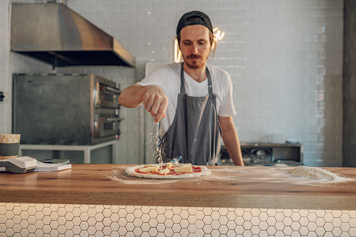 Man pizza master prepares pizza according to customer requests. Process of placing ingredients on a pizza. Pizza place and tasty food concept. Focus on a man sprinkling cheese.
