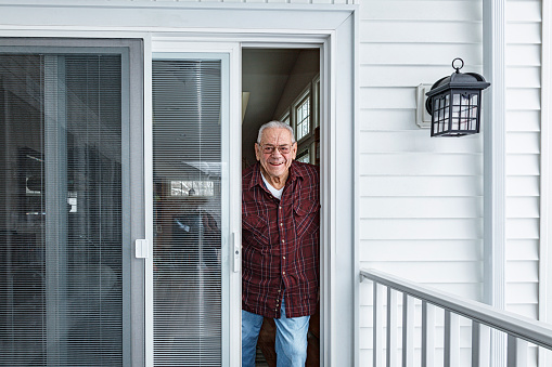 A real life, real person elderly senior adult man is standing in the doorway of his home front door looking out at the camera with a cheerful smile.