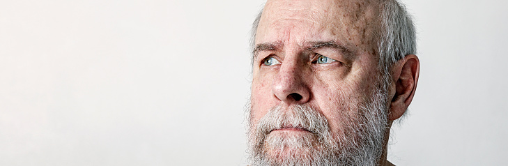 Envelope shape composition cropped portrait of a somewhat scruffy, green eyed, tangled hair, gray bearded senior adult man looking upwards away from the camera with a curious or questioning facial expression. He has some dry, peeling skin above his temple.