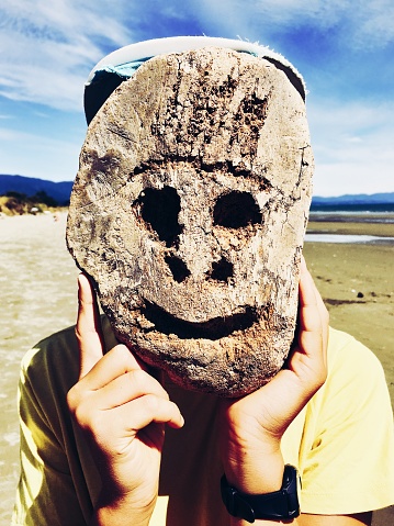 A boy holds up his hand carved wooden mask in front of his face at the beach in summer. The mask is carved from a tree fern log piece of driftwood.