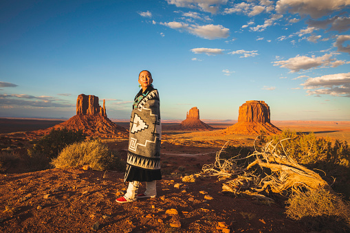 A Native American woman dressed in traditional Navajo clothing, poses in the landscape of Monument Valley, on the border of Utah and Arizona.