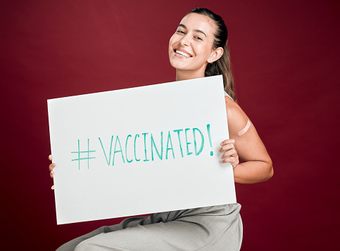 Mixed race covid vaccinated woman showing plaster on arm and holding poster. Portrait of smiling hispanic woman isolated against red studio background with copyspace. Promoting corona vaccine on sign