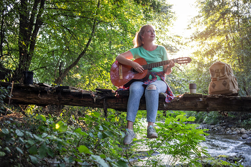 One young woman is staying in nature by the river and playing the guitar, she has created an atmosphere for enjoyment and relaxation.