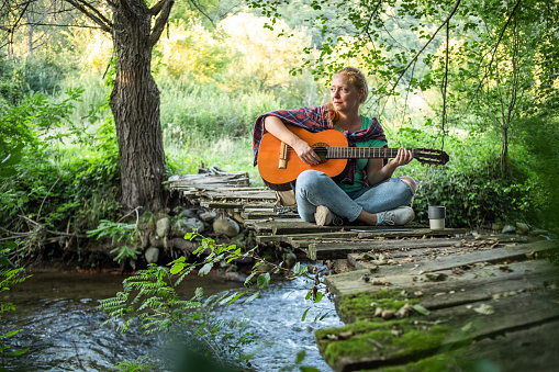 One young woman is staying in nature by the river and playing the guitar, she has created an atmosphere for enjoyment and relaxation.