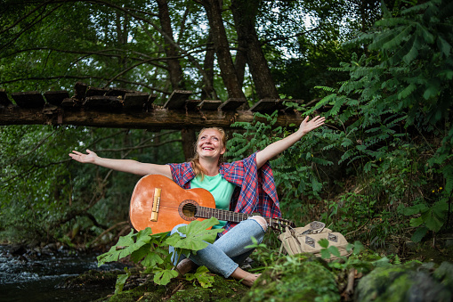 One young woman is staying in nature by the river and playing the guitar, she has created an atmosphere for enjoyment and relaxation.