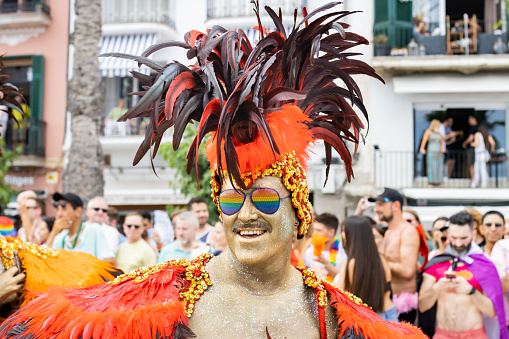Sitges, Spain - June 12, 2022: Smiling man with his face painted with a colorful feathered hat and glasses celebrating the gay pride event in Sitges, Spain.