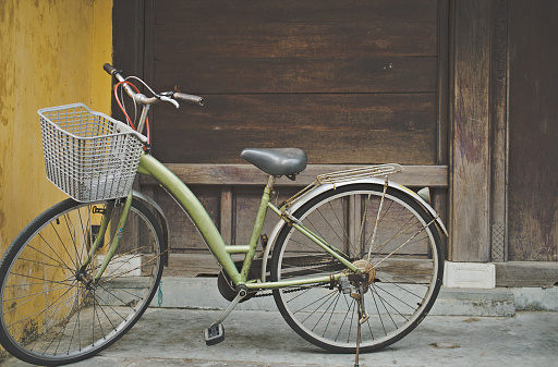 green bike with a basket parked in front of an old wooden facade building in the historical center of Hoi An city. Bicycle is the best way of discovering the old quarter and its beautiful weathered houses.