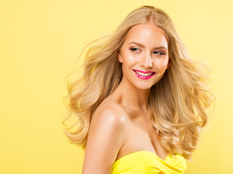 Blond Woman with Long Curly Hairstyle. Blonde Fashion Girl with Summer Face Makeup enjoying Sun. Beauty Model Happy smiling Portrait over bright Yellow Studio background