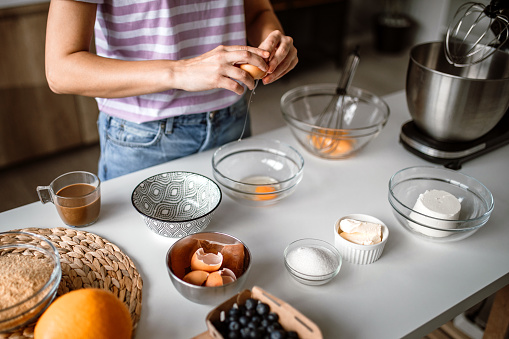 Mid adult woman making cheesecake in home kitchen, cracking an egg for preparing a cake batter.