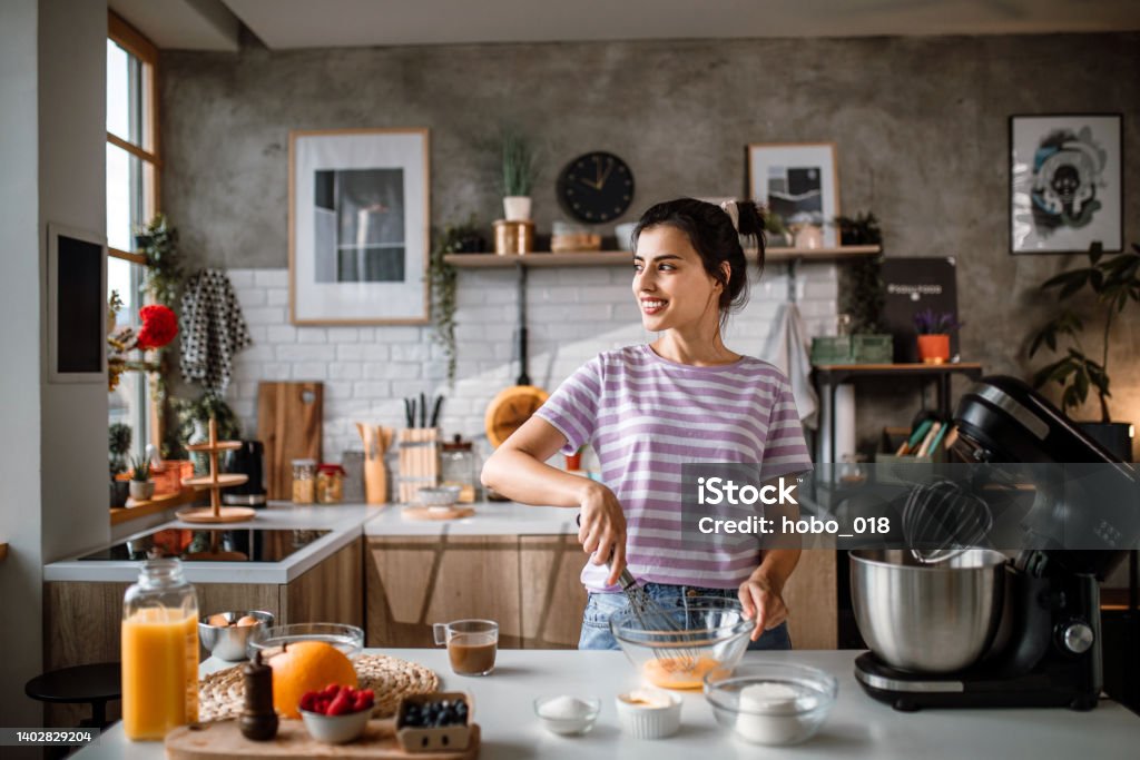 Hand whisking eggs is first step for best cakes. Mid adult woman making cheesecake in home kitchen. Whisking cake mixture in a glass bowl, looking the through window. Cooking Stock Photo