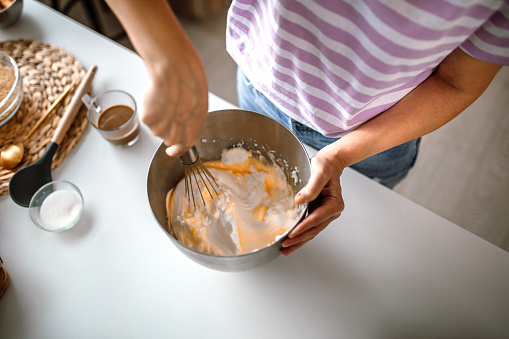 Mid adult woman making cheesecake in home kitchen. Whisking egg yolks and egg whites mixture in inox bowl.