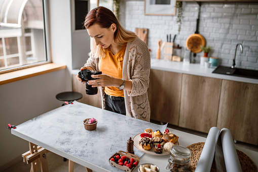 Young female photographer standing over cakes on the table, holding camera, photographing cakes and cookies for her new vlog.