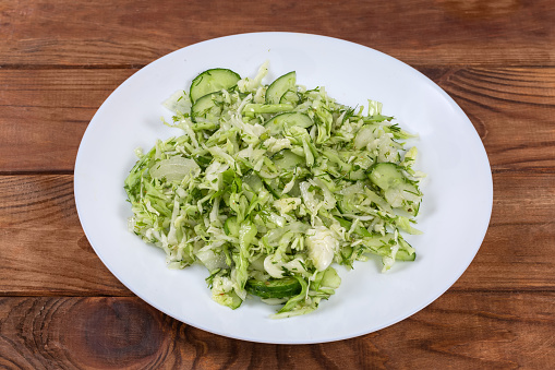 Green spring vegetable salad made of sliced fresh young cabbage, cucumbers, chopped onion and dill on a white dish on an old rustic table