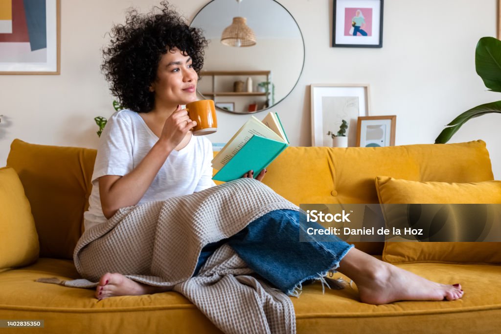 ペンシブなリラックスしたアフリカ系アメリカ人の女性は、ソファに座ってコーヒーを飲みながら、家で本を読んでいます。スペースをコピーします。 - リラクゼーションのロイヤリティフリーストックフォト