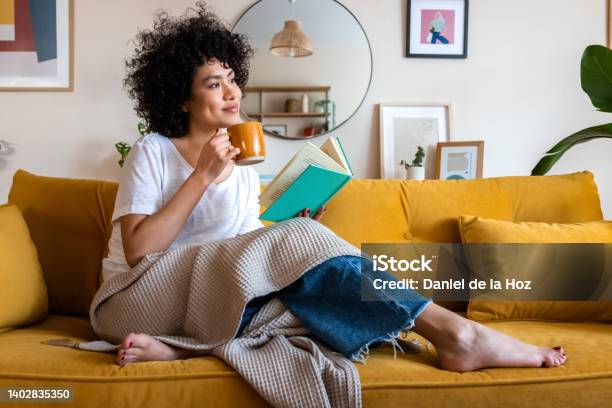 ペンシブなリラックスしたアフリカ系アメリカ人の女性はソファに座ってコーヒーを飲みながら家で本を読んでいますスペースをコピーします - リラクゼーションのストックフォトや画像を多数ご用意