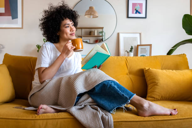 pensive relaxed african american woman reading a book at home, drinking coffee sitting on the couch. copy space. - vrije tijd fotos stockfoto's en -beelden