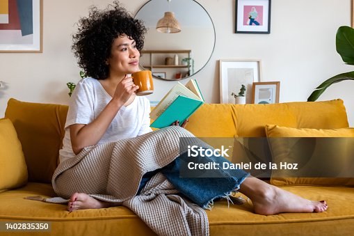 istock Pensive relaxed African american woman reading a book at home, drinking coffee sitting on the couch. Copy space. 1402835350
