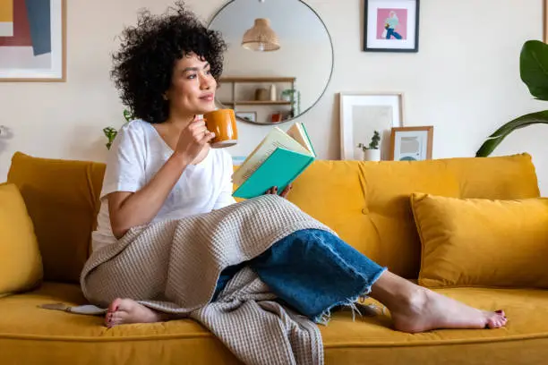 Photo of Pensive relaxed African american woman reading a book at home, drinking coffee sitting on the couch. Copy space.