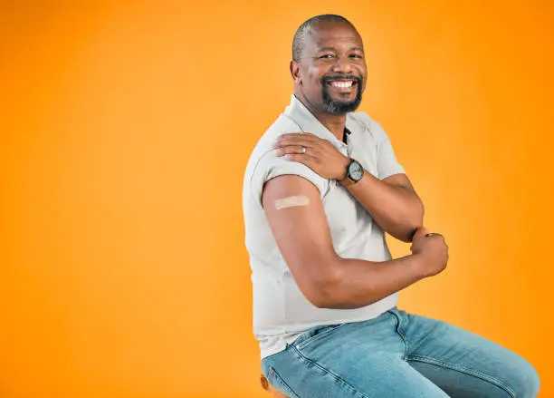 Photo of African american covid vaccinated man showing plaster on arm and smiling. Portrait of black man isolated against yellow studio background with copyspace. Protected from corona with vaccine injection