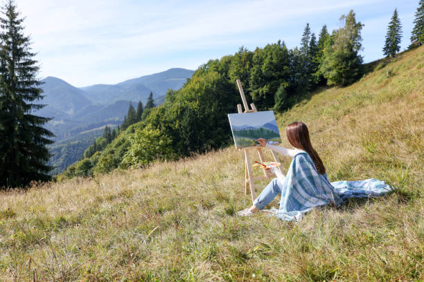 istock Young woman drawing on easel in mountains, back view. Space for text 1402841084